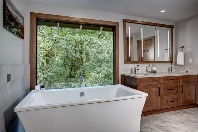 Primary bathroom with a freestanding tub under a large window onto the trees and a wood vanity